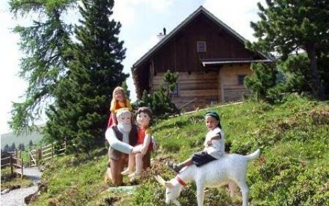 Drei Kinder posieren neben lebensgroßen Statuen von Figuren und einer Ziege auf einem grasbewachsenen Hügel mit einer Holzhütte und Bäumen im Hintergrund.