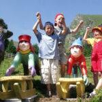 Drei Kinder stehen zwischen bunten Zwergenstatuen und Holzschlitten im Freien und feiern mit erhobenen Armen; ein weiteres Kind und eine Zwergenstatue stehen in der Nähe, mit Bergen im Hintergrund.