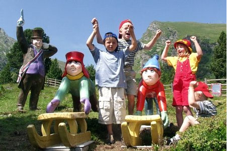 Drei Kinder stehen zwischen bunten Zwergenstatuen und Holzschlitten im Freien und feiern mit erhobenen Armen; ein weiteres Kind und eine Zwergenstatue stehen in der Nähe, mit Bergen im Hintergrund.
