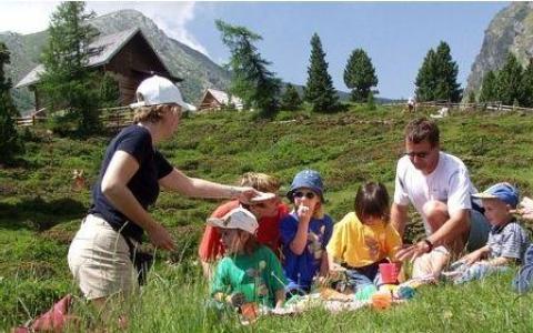 Eine Gruppe von Erwachsenen und Kindern macht ein Picknick auf einem grasbewachsenen Hügel mit Bäumen, Bergen und einer Hütte im Hintergrund.