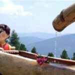 Statue eines Kindes neben einem hölzernen Wassertrog mit einem Blumenstrauß, aus dessen Ausguss Wasser fließt; im Hintergrund sind Berge und Bäume zu sehen.