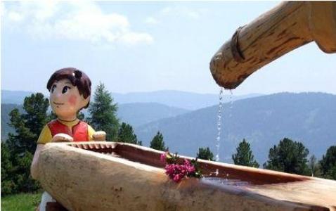 Statue eines Kindes neben einem hölzernen Wassertrog mit einem Blumenstrauß, aus dessen Ausguss Wasser fließt; im Hintergrund sind Berge und Bäume zu sehen.