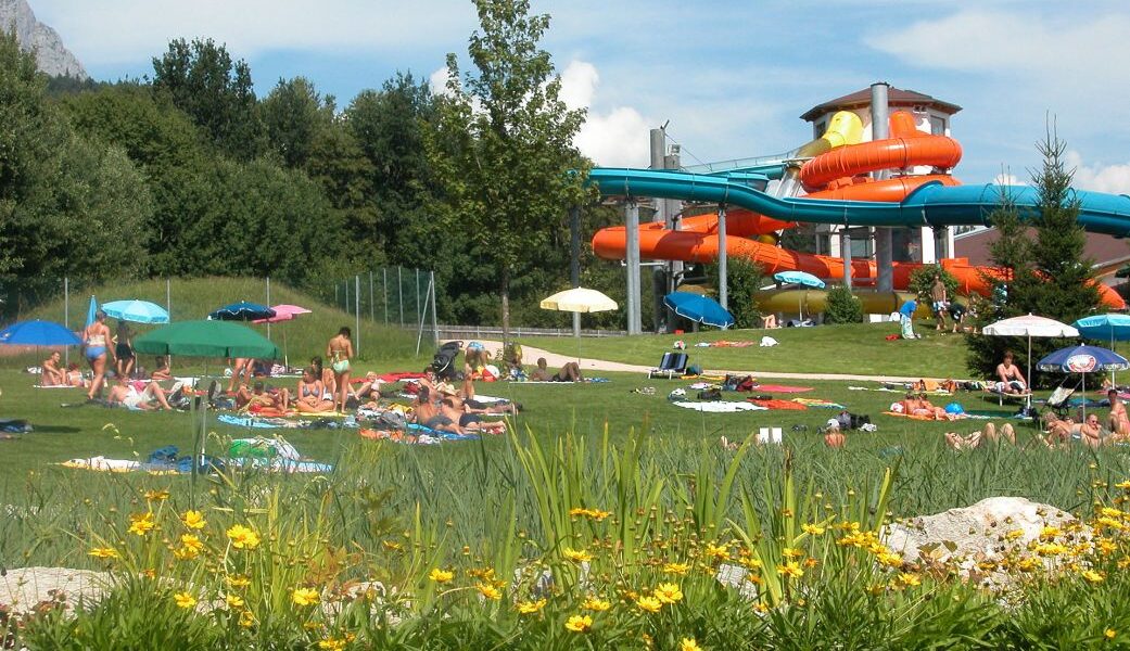 Menschen entspannen sich auf einer Wiese mit Handtüchern und Sonnenschirmen in der Nähe eines Wasserparks mit orangefarbenen und blauen Wasserrutschen, umgeben von Bäumen und Blumen.