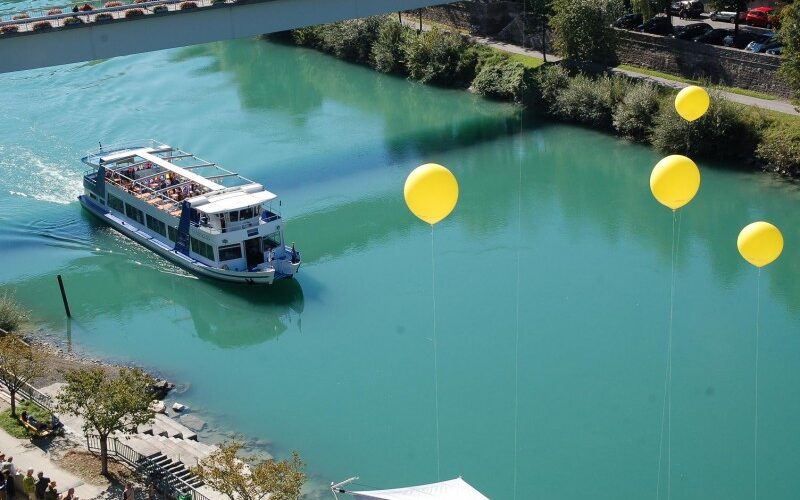 Ein Passagierboot fährt auf einem türkisfarbenen Fluss unter einer Brücke hindurch, in dessen Nähe mehrere gelbe Luftballons schwimmen und Bäume das Ufer säumen.