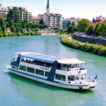 Ein weißes Flussboot fährt auf einem grünlichen Fluss mit städtischen Gebäuden, Bäumen und einem Kirchturm im Hintergrund unter einem klaren Himmel.