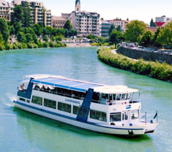 Ein weißes Flussboot fährt auf einem grünlichen Fluss mit städtischen Gebäuden, Bäumen und einem Kirchturm im Hintergrund unter einem klaren Himmel.