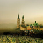 Ein großes historisches Gebäude mit grünen Kuppeln und Zwillingstürmen steht hinter einem Weinberg, umgeben von nebligen Hügeln und einem dunstigen Himmel.