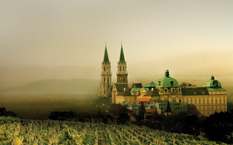 Ein großes historisches Gebäude mit grünen Kuppeln und Zwillingstürmen steht hinter einem Weinberg, umgeben von nebligen Hügeln und einem dunstigen Himmel.