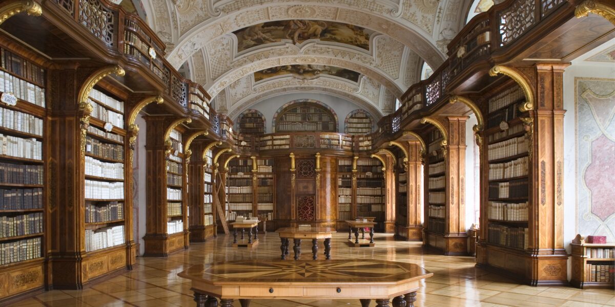 Eine prächtige, verschnörkelte Bibliothek mit hohen Holzregalen, gewölbten Deckenfresken und einem runden Holztisch in der Mitte des polierten Bodens.