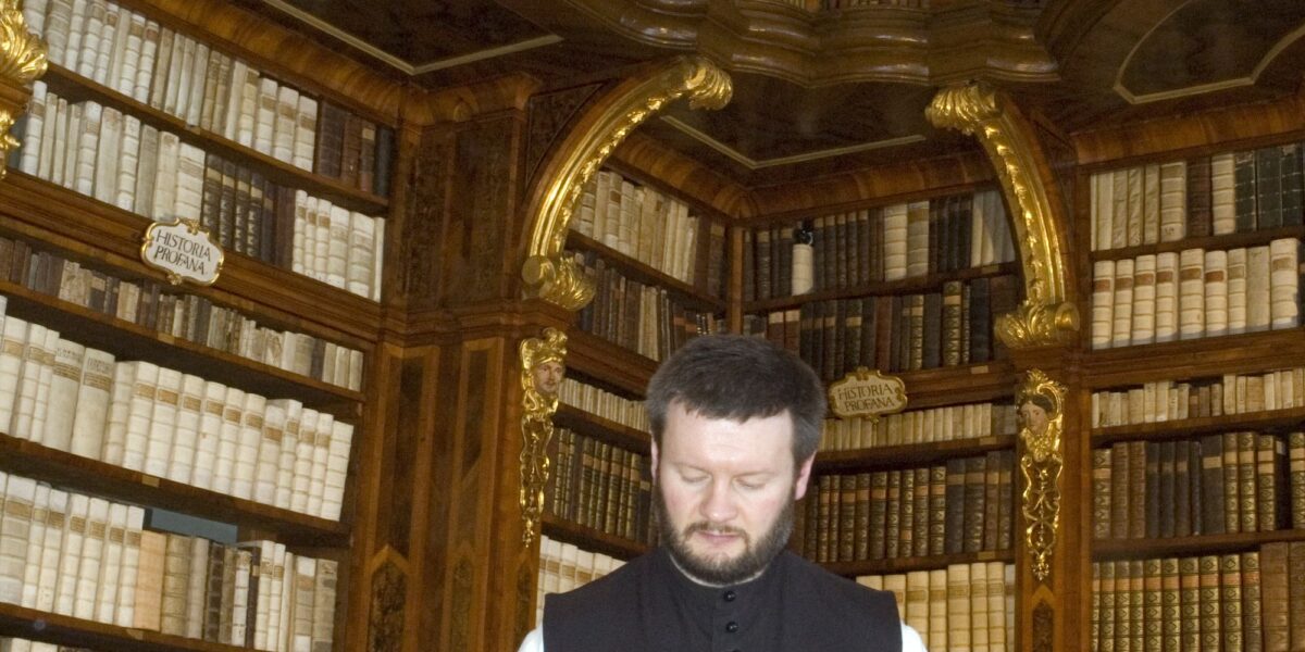 Ein Mann in einer schwarz-weißen Weste liest ein großes Buch an einem Tisch in einer kunstvollen Bibliothek mit hohen Regalen und gewölbten Decken, die mit Büchern gefüllt sind.
