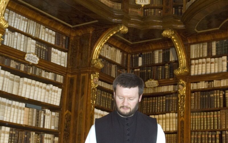 Ein Mann in einer schwarz-weißen Weste liest ein großes Buch an einem Tisch in einer kunstvollen Bibliothek mit hohen Regalen und gewölbten Decken, die mit Büchern gefüllt sind.