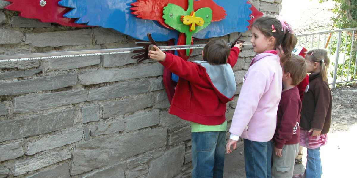 Vier Kinder stehen in einer Reihe und messen ihre Armspannweite anhand eines bunten, vogelförmigen Wanddisplays, das an einer Steinmauer angebracht ist.