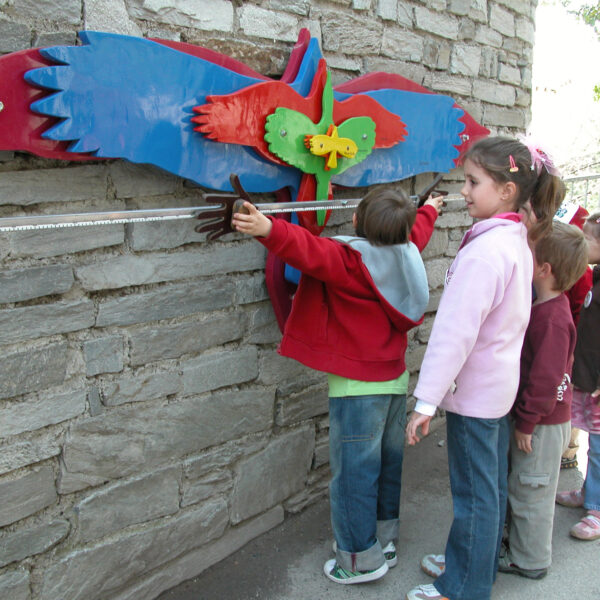Vier Kinder stehen in einer Reihe und messen ihre Armspannweite anhand eines bunten, vogelförmigen Wanddisplays, das an einer Steinmauer angebracht ist.