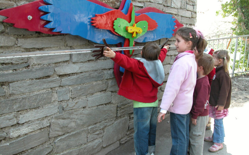 Vier Kinder stehen in einer Reihe und messen ihre Armspannweite anhand eines bunten, vogelförmigen Wanddisplays, das an einer Steinmauer angebracht ist.