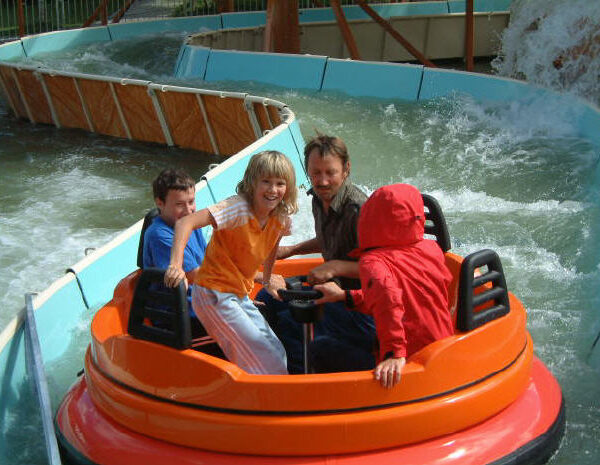 Vier Personen fahren auf einem runden orangefarbenen Floß auf einer Wasserbahn, im Hintergrund sind spritzendes Wasser und gebogene Barrieren zu sehen.