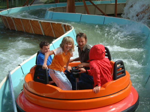 Vier Personen fahren auf einem runden orangefarbenen Floß auf einer Wasserbahn, im Hintergrund sind spritzendes Wasser und gebogene Barrieren zu sehen.