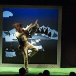 Ein Darsteller auf der Bühne hält ein Schaf vor einem cartoonhaften Hintergrund, der Berge, Bäume und einen mondbeschienenen Himmel zeigt, während er auf grünem Rasen steht.