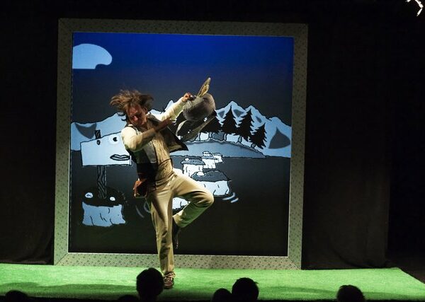 Ein Darsteller auf der Bühne hält ein Schaf vor einem cartoonhaften Hintergrund, der Berge, Bäume und einen mondbeschienenen Himmel zeigt, während er auf grünem Rasen steht.