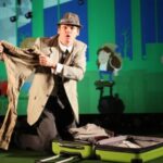 Ein Mann in Anzug und Hut kniet neben einem offenen Koffer und hält eine Hose hoch, während hinter ihm eine Reiseszene im Cartoon-Stil projiziert wird.