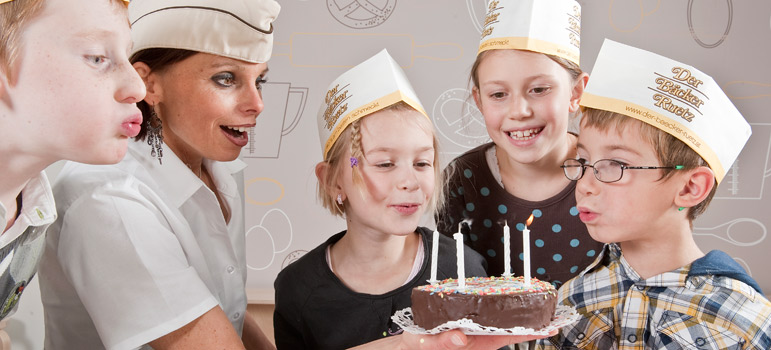 Vier Kinder und ein Erwachsener mit Papierhüten haben sich um einen kleinen Schokoladenkuchen mit brennenden Kerzen versammelt und bereiten sich darauf vor, diese gemeinsam auszublasen.