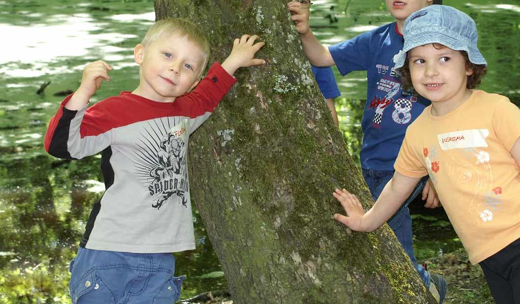 Vier junge Kinder stehen im Freien an einem Baum, wobei zwei von ihnen den Baumstamm berühren. Sie sind leger gekleidet und scheinen sich in einem Park oder Naturgebiet aufzuhalten.