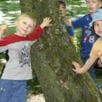 Vier junge Kinder stehen im Freien an einem Baum, wobei zwei von ihnen den Baumstamm berühren. Sie sind leger gekleidet und scheinen sich in einem Park oder Naturgebiet aufzuhalten.