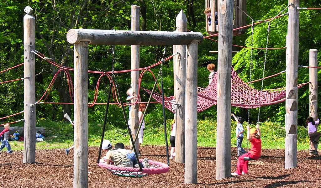 Kinder spielen auf einem von Bäumen umgebenen Spielplatz auf einer Kletterstruktur aus Holz und Seilen.
