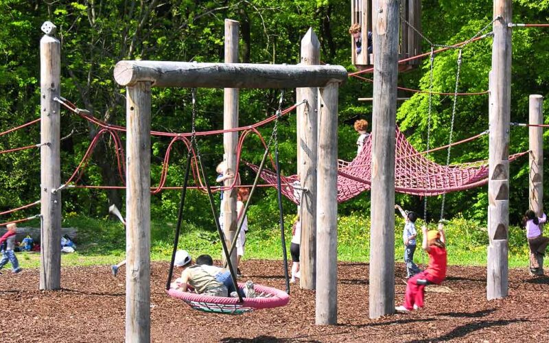 Kinder spielen auf einem von Bäumen umgebenen Spielplatz auf einer Kletterstruktur aus Holz und Seilen.