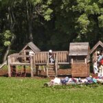 Kinder spielen auf einem hölzernen Spielplatz, der von Bäumen umgeben ist, während im Vordergrund Rucksäcke und Jacken auf dem Gras liegen.