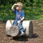 Ein kleiner Junge sitzt auf einem Holzblock in Form eines Tieres und schützt seine Augen vor der Sonne, während er in einem Park in die Ferne schaut.