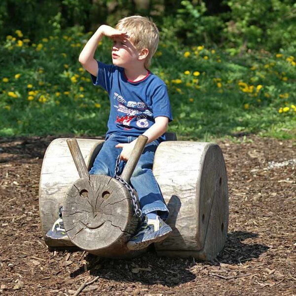 Ein kleiner Junge sitzt auf einem Holzblock in Form eines Tieres und schützt seine Augen vor der Sonne, während er in einem Park in die Ferne schaut.