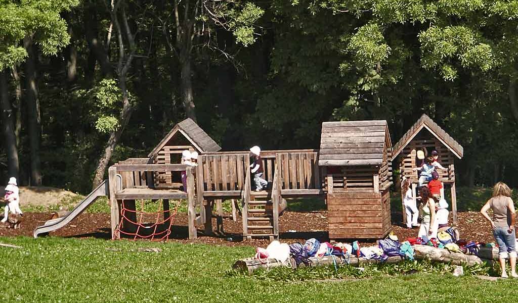 Kinder spielen auf einem hölzernen Spielplatz mit einer Rutsche und einem Kletternetz, während Taschen und Gegenstände auf dem Gras im Vordergrund verstreut sind. Der Hintergrund ist von Bäumen gesäumt.
