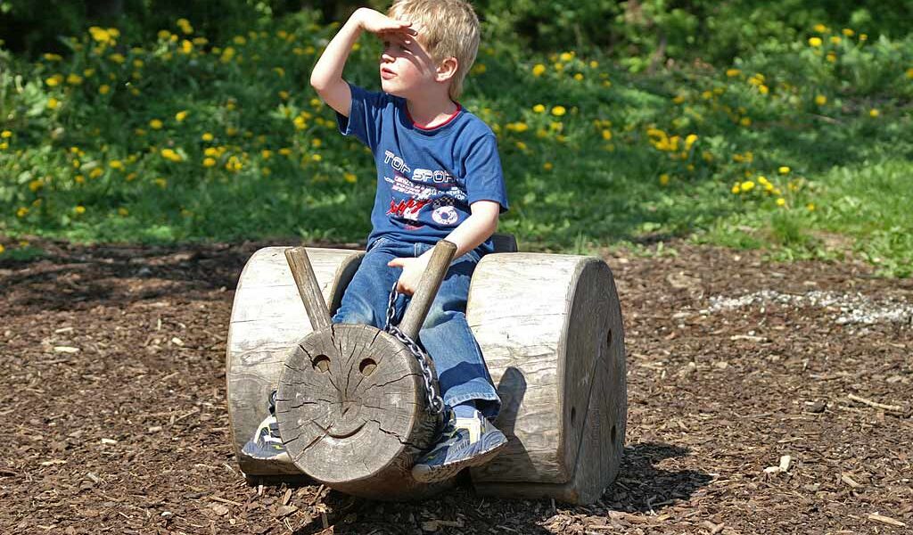 Ein kleiner Junge sitzt auf einem hölzernen Spielplatz, der die Form eines Gesichts hat, und schützt seine Augen mit der Hand vor der Sonne.