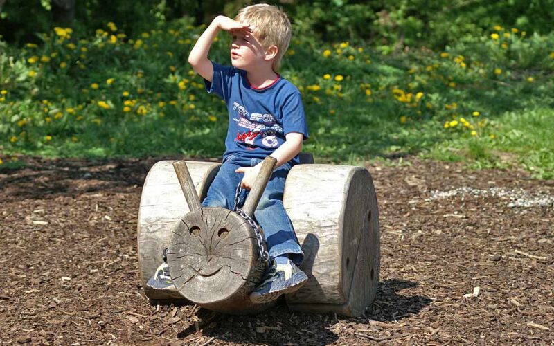 Ein kleiner Junge sitzt auf einem hölzernen Spielplatz, der die Form eines Gesichts hat, und schützt seine Augen mit der Hand vor der Sonne.