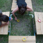 Zwei Personen liegen auf Holzbänken im Freien, halten jeweils eine Rolle Klebeband in der Hand und arbeiten an einem Projekt auf der Wiese zwischen den Bänken.
