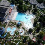Luftaufnahme eines öffentlichen Freibadkomplexes mit Wasserrutschen, Sonnenanbetern auf der Wiese und einem nahe gelegenen Basketballplatz.
