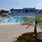 Freibadkomplex mit Menschen im Wasser, einem Rettungsschwimmerturm, blau-weißem Sitzbereich und einer Grünanlage mit Blumen im Vordergrund.
