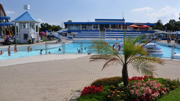 Freibadkomplex mit Menschen im Wasser, einem Rettungsschwimmerturm, blau-weißem Sitzbereich und einer Grünanlage mit Blumen im Vordergrund.