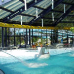 Hallenbad mit Wasserdüsen, Wasserfällen und einem transparenten Schrägdach; eine Person schwimmt, und rund um den Poolbereich sind Pflanzen zu sehen.