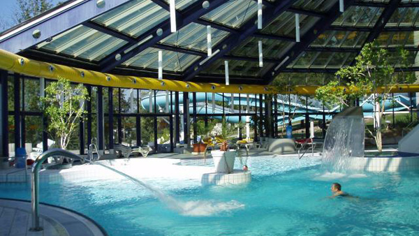 Hallenbad mit Wasserdüsen, Wasserfällen und einem transparenten Schrägdach; eine Person schwimmt, und rund um den Poolbereich sind Pflanzen zu sehen.