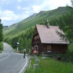 Ein Mann steht in der Nähe eines kleinen Holzhauses an einer kurvigen Bergstraße, umgeben von grünen Hügeln und Bäumen unter einem teilweise bewölkten Himmel.