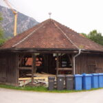 Ein achteckiges Holzgebäude mit Ziegeldach, umgeben von Bäumen, vor dem blaue und schwarze Mülltonnen aufgereiht sind und in dem Bauschutt liegt.