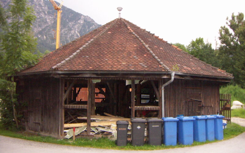 Ein achteckiges Holzgebäude mit Ziegeldach, umgeben von Bäumen, vor dem blaue und schwarze Mülltonnen aufgereiht sind und in dem Bauschutt liegt.