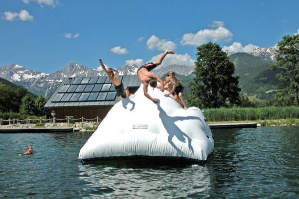 Vier Personen spielen und springen von einem großen Schlauchboot, das auf einem See schwimmt, mit Bergen, Bäumen und einem Gebäude im Hintergrund unter einem sonnigen Himmel.