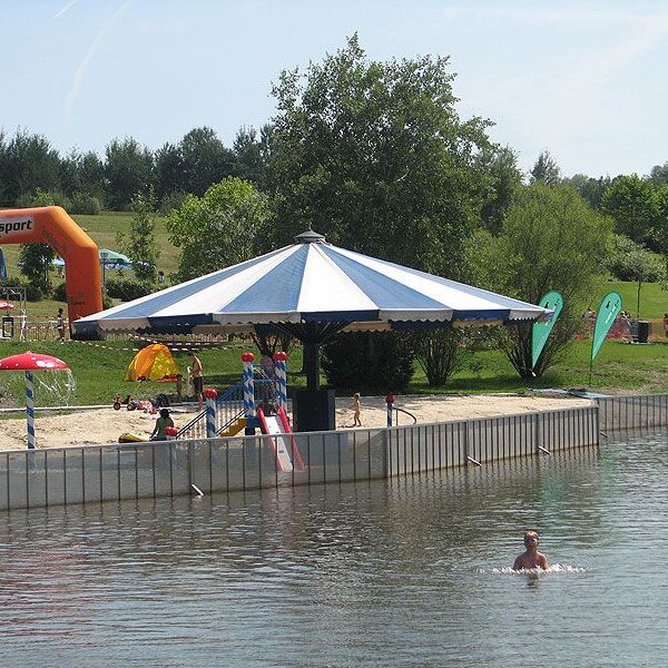 Eine Person schwimmt in einem Teich in der Nähe eines Stegs mit einer großen Schirmkonstruktion; Kinder und Familien spielen und entspannen sich auf der Wiese im Hintergrund.