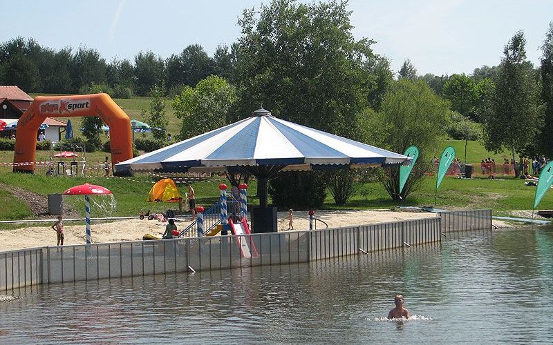 Eine Person schwimmt in einem Teich in der Nähe eines Stegs mit einer großen Schirmkonstruktion; Kinder und Familien spielen und entspannen sich auf der Wiese im Hintergrund.