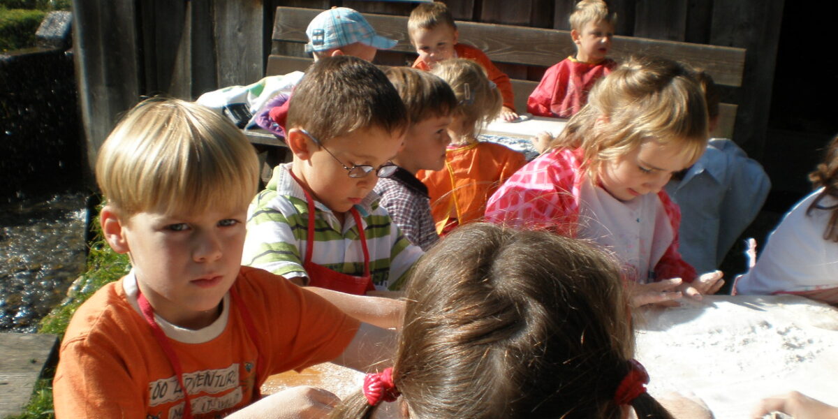 Eine Gruppe kleiner Kinder sitzt im Freien an einem Tisch und beschäftigt sich mit weißem, pulverförmigem Material.