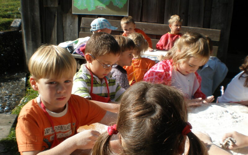 Eine Gruppe kleiner Kinder sitzt im Freien an einem Tisch und beschäftigt sich mit weißem, pulverförmigem Material.