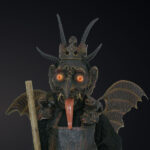 Eine Figur, die eine gehörnte, dämonische Maske mit glühenden Augen, fledermausähnlichen Flügeln und einer Krone trägt und einen Holzstab in der Hand hält, steht vor einem dunklen Hintergrund.