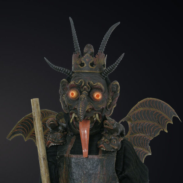 Eine Figur, die eine gehörnte, dämonische Maske mit glühenden Augen, fledermausähnlichen Flügeln und einer Krone trägt und einen Holzstab in der Hand hält, steht vor einem dunklen Hintergrund.